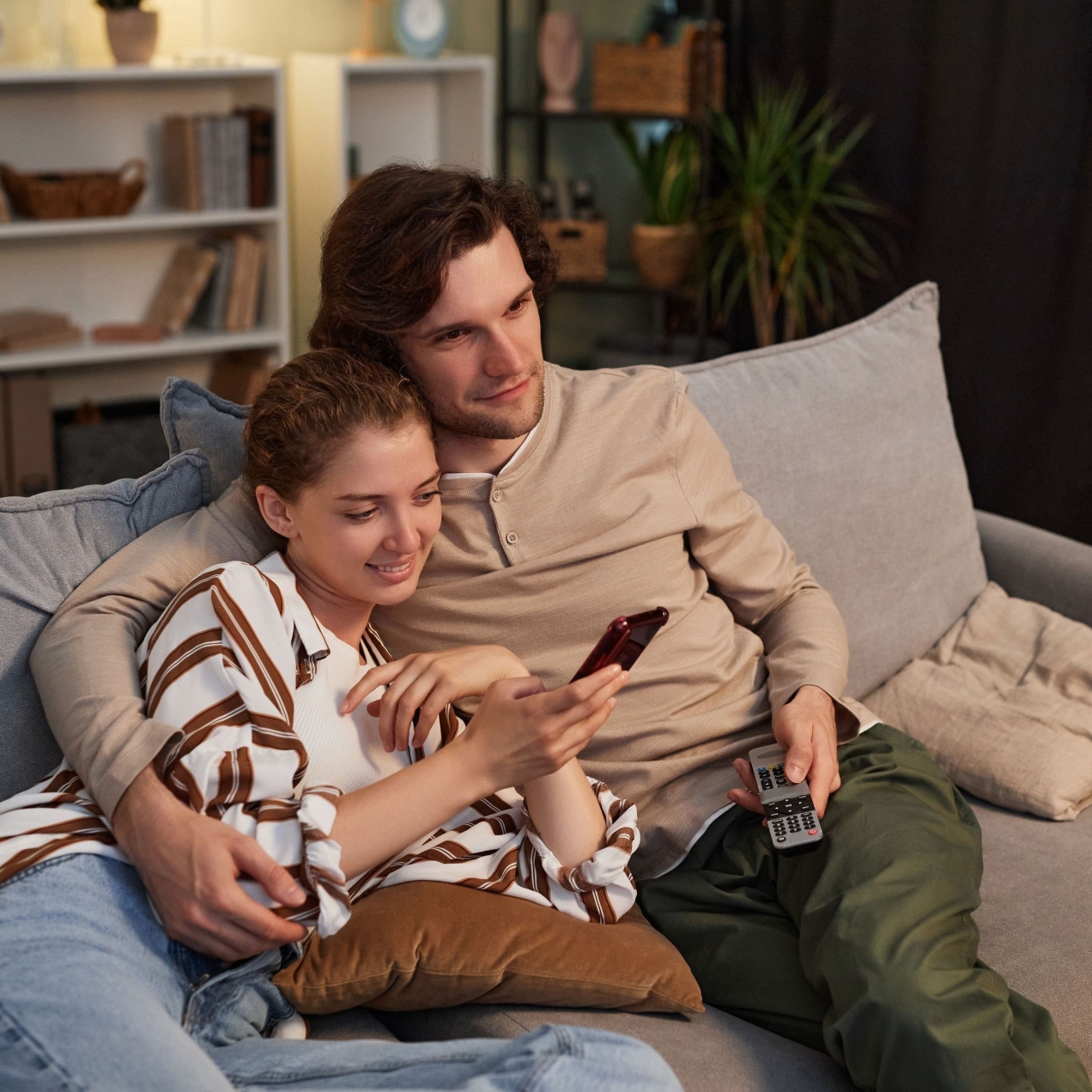 A white man with short dark hair wearing a light top and dark trousers holding a TV remote control sits on a sofa with his arm round a white woman who is wearing a striped top and light jeans and is looking at a mobile phone
